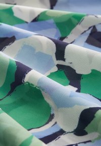 Tissu texturé présentant un motif abstrait avec des formes superposées vertes, bleues et noires sur un fond clair, mettant en valeur des couleurs vives.