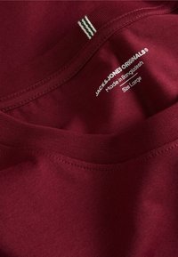T-shirt en coton bordeaux avec col rond, comportant une étiquette indiquant la marque, le pays de fabrication et la taille. Deux bandes blanches accentuent le col.