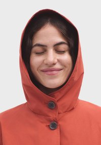 Giacca con cappuccio rosso dal tessuto morbido, caratterizzata da due bottoni scuri e un design semplice. Il cappuccio è aderente, incorniciando il viso.