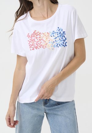 Kvinde iført hvid t-shirt med et pixeleret gradientdesign i rødt, gult og blåt på brystet, kombineret med lyseblå jeans.
