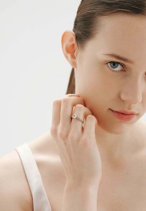 Mujer joven con ojos azules que se toca la cara, lleva una blusa blanca y un anillo de plata con una piedra preciosa transparente en el dedo.