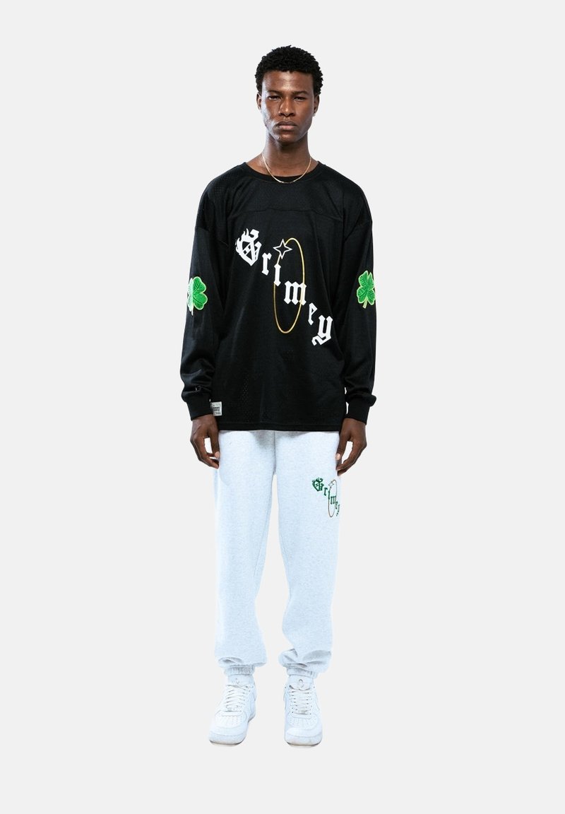 Camisa negra de manga larga con gráficos en blanco y verde, combinada con pantalones de chándal gris claro con logo verde y zapatillas blancas.