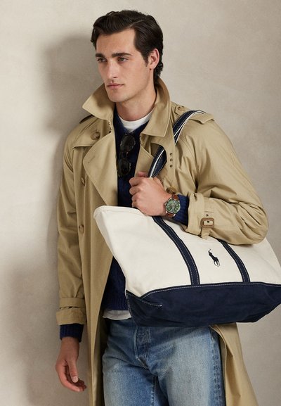 Bărbat purtând o trenci bej, pulover albastru, blugi și ceas verde, ținând o geantă mare albă și bleumarin cu logo-ul unui cal negru.