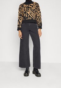 Person in einem braun-schwarzen Tiermuster-Pullover, schwarzen weit geschnittenen Cropped-Jeans und schwarzen Schnürstiefeln vor einem einfarbigen Hintergrund.