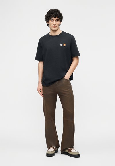 Νεαρός με σγουρά μαλλιά φοράει μαύρο μπλουζάκι με υφασμάτινα σχέδια ζώων, καφέ καμπάνα παντελόνι και μπεζ μοκασίνια, στέκεται με το ένα χέρι στην τσέπη.