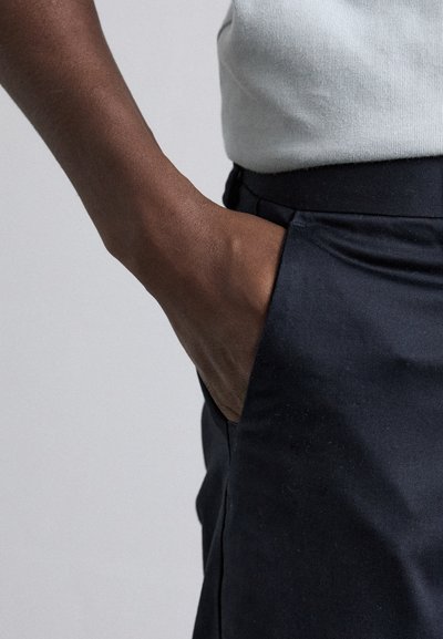 Pantalones negros de tela suave, con un bolsillo lateral y un diseño ajustado. Una mano descansando en el bolsillo añade detalle.