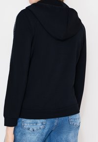 Personne portant un sweat à capuche noir avec la capuche baissée et un jean bleu délavé, photographiée de dos sur un fond blanc.