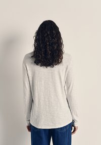 Svetlošedá košeľa s dlhým rukávom vyrobená z textúrovaného materiálu, s okrúhlym lemom a voľným strihom, v kombinácii s modrými rifľami.