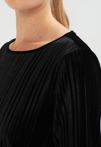 Schwarzes Samtoberteil mit vertikalen Falten, Rundhalsausschnitt und langen Ärmeln. Der Stoff hat eine weiche Textur und einen dezenten Glanz.