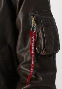 Jaqueta bomber de couro castanho com um bolso com fecho, superfície texturizada e uma etiqueta de tecido vermelha com a inscrição "REMOVER ANTES DO VOO".