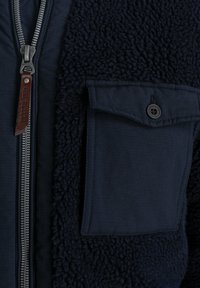 Navy blue jacket with a textured fleece body and smooth fabric sleeve. Features a metal zipper and a chest pocket with a button.