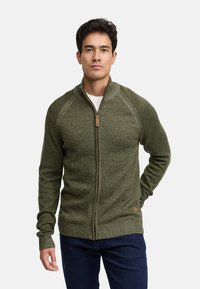 Pull en zip vert avec un motif de maille texturé, des poignets et un ourlet côtelés, présentant une languette en cuir sur la fermeture éclair et un petit détail de logo.