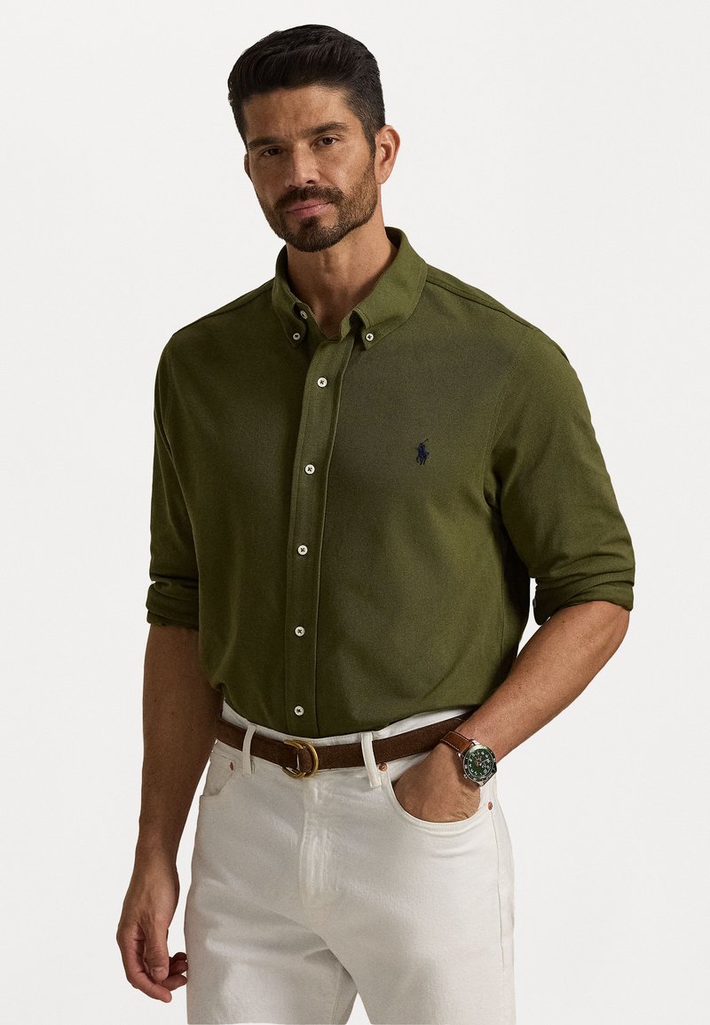 Camisa verde-azeitona abotoada com colarinho, apresentando um pequeno logótipo azul-marinho no peito, usada com calças brancas e um cinto castanho.