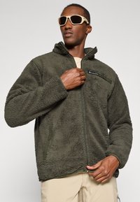 Fleece-Jacke in Dunkelgrün, mit hohem Kragen, Reißverschluss, seitlichen Taschen und einem Logo-Patch auf der linken Brust. Weiche Textur.