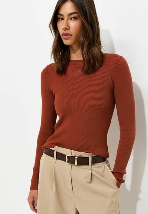 Vrouw draagt een aansluitende roestkleurige geribde trui en beige hoge taille broek met een donkerbruine riem, staand tegen een witte achtergrond.