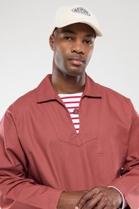 Rode katoenen shirt met een brede kraag en V-hals, gelaagd over een wit met rood gestreept shirt. De persoon draagt een lichtbeige pet.
