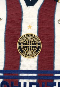 Vêtement de sport en maille blanche avec des rayures verticales bordeaux, un patch circulaire doré et noir « Prohibited Sportswear Berlin, Germany » et une étoile dorée au-dessus.