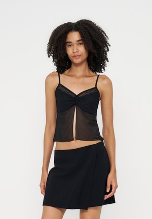 Frau mit lockigem Haar, die ein schwarzes durchsichtiges Camisole-Oberteil und einen schwarzen Rock trägt, steht vor einem schlichten hellen Hintergrund.