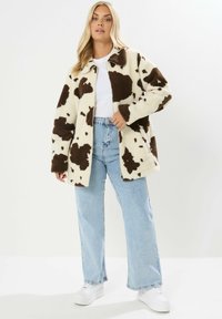 Fleece jack met een wit en bruin koeienpatroon, met kraagontwerp, relaxte pasvorm, gecombineerd met lichtblauwe jeans en witte sneakers.