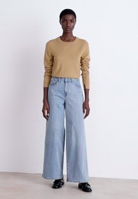 Suéter beige de manga larga combinado con unos jeans de pierna ancha y cintura alta de color azul claro. El modelo lleva zapatos negros con una textura brillante.