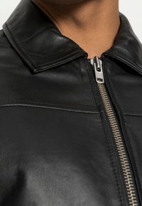 Primo piano di una giacca di pelle nera con cerniera chiusa, che mostra il colletto e la cerniera argentata su uno sfondo di collo simile alla pelle.