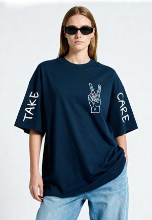 Vrouw met zwarte zonnebril en een oversized navy t-shirt met vredeshand symbool en de woorden "Take Care" op de mouwen en spijkerbroek.