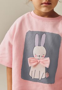 T-shirt en coton rose avec un graphique de lapin gris aux oreilles à carreaux violets, un nœud rose et un accent floral sur le côté du lapin.