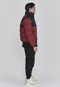 Chaqueta tipo puffer en negro y burdeos con cuello alto, cierre de cremallera en la parte frontal y patrón de acolchado rectangular. Combinada con pantalones cargo negros y zapatillas deportivas.
