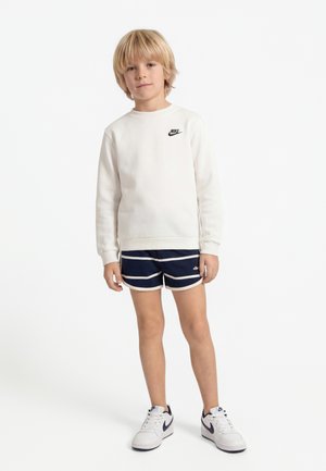 Blond boy standing, wearing a white Nike sweatshirt, navy striped shorts, and white Nike sneakers on a white background.