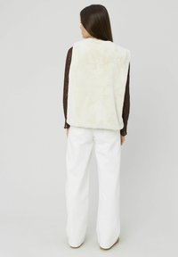 Gilet en fausse fourrure écru avec une texture douce, présentant un col rond et des côtés ouverts, porté sur un top en dentelle foncée et un pantalon large blanc.