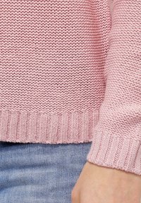 Pull en tricot rose avec un ourlet et des poignets côtelés, présentant un motif texturé et une coupe décontractée. Le matériau est doux et respirant.