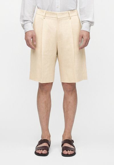 Homme portant un short plissé beige au genou, une chemise rayée claire et des sandales marron foncé, debout sur un fond blanc uni.
