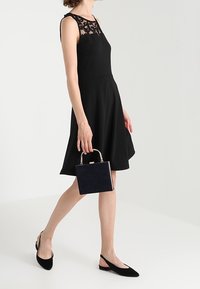 Czarna sukienka bez rękawów z koronkowym dekoltem, rozkloszowana spódnica; modelka trzyma małą granatową torebkę na złotym zapięciu, w połączeniu z czarnymi czółenkami.