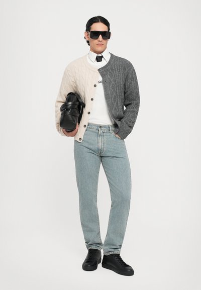 Tvåfärgad kabelstickad cardigan i kräm och grått, vit skjorta, svart slips, ljusblå jeans och svarta sneakers med strukturerad yta.