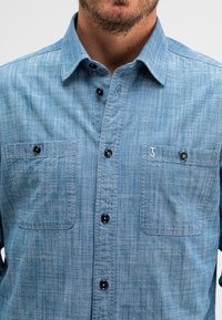 Lichtblauwe knoopshirts met twee borstzakken, zwarte knopen en een subtiele textuurstof. Bevat een geborduurd logo op de zak.