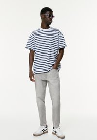 Mann mit dunkler Hautfarbe, der eine Sonnenbrille trägt, ein weiß-blau gestreiftes T-Shirt, hellgraue Jeans und weiße Sneaker mit schwarzen Details.