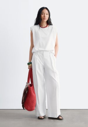Femme portant un haut blanc sans manches et un pantalon large, tenant un sac en tricot rouge avec une écharpe marron, et portant des sandales et des bijoux.