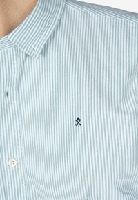Camisa de rayas azul claro con cuello abotonado, hecha de una tela que presenta finas líneas verticales y un pequeño detalle bordado en azul marino.