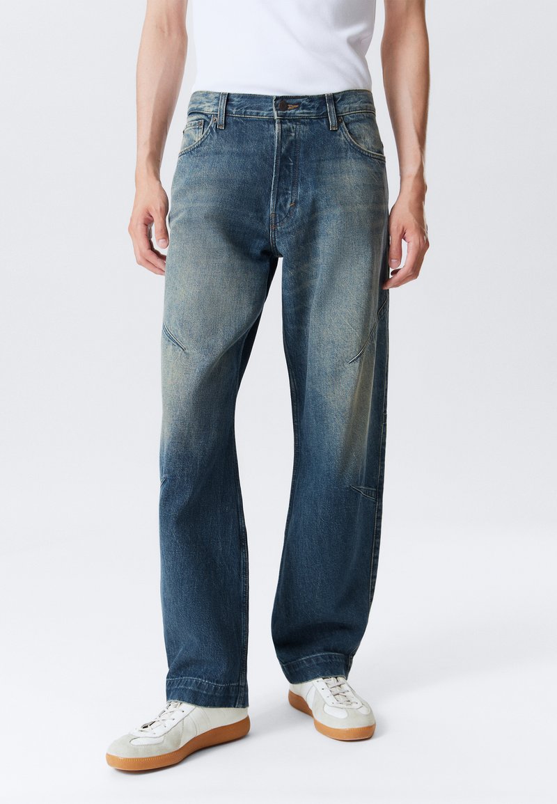 Henkilö, joka on pukeutunut haalistuneisiin sinisiin suoriin farkkuihin, valkoisiin kumipohjallisiin tennareihin ja valkoiseen hihattomaan paitaan, seisoo tasaisella taustalla.