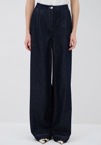 Leveleppa tummasta farkkukankaasta, jossa on korkea vyötärö, etupleatit ja yksittäinen nappisuljenta. Ominaisuudet: valkoinen ompelu ja käänteet lahkeissa.