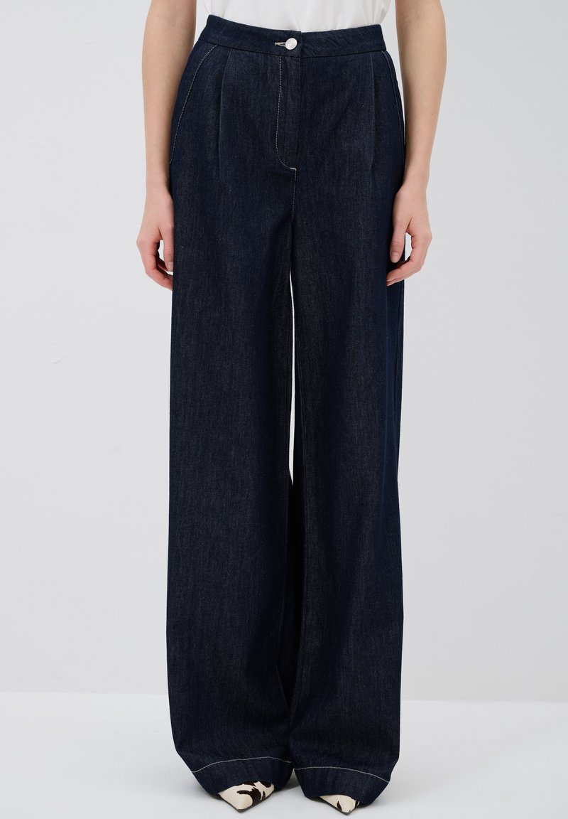 Leveleppa tummasta farkkukankaasta, jossa on korkea vyötärö, etupleatit ja yksittäinen nappisuljenta. Ominaisuudet: valkoinen ompelu ja käänteet lahkeissa.