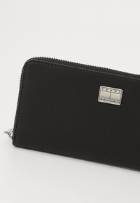Portafoglio in pelle nera con finitura texture, dotato di una placca logo in argento e di una chiusura a zip lungo il perimetro.