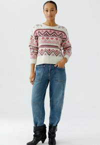 Gebreide trui met een crèmekleurige basis en geometrische patronen in rood, roze en zwart, gecombineerd met losse blauwe denim jeans en zwarte laarzen tot de knie.