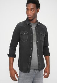 Camicia di denim nera con chiusura a bottoni sul davanti, due tasche sul petto e maniche arrotolate. Indossata sopra una t-shirt grigia con stampa, abbinata a jeans grigi.