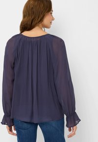 Blusa a maniche lunghe in tessuto trasparente blu scuro, con retro plissettato, polsini elasticizzati e scollatura tonda. Abbinata a jeans blu.