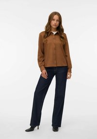 Braunes Hemd mit langen Ärmeln, aus weichem Stoff, kombiniert mit dunkelblauen Denim-Hosen im weiten Schnitt und schwarzen, hochhackigen Stiefeletten.