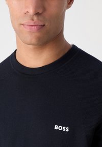 Μπλε πουλόβερ crewneck από υφασμάτινη πλέξη με λευκό λογότυπο "BOSS" κεντημένο στο στήθος. Διαθέτει σχέδιο με ραγλανικά μανίκια.