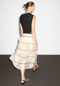 Femme marchant à l'intérieur portant un haut sans manches noir, une jupe midi crème à rayures horizontales, des sandales noires et des boucles d'oreilles remarquables, les cheveux relevés en chignon.