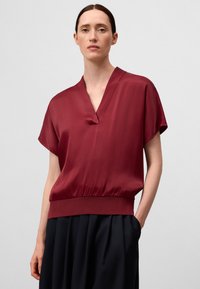 Top in seta bordeaux con scollatura a V, maniche corte e orlo elasticizzato. Texture liscia e vestibilità comoda, abbinato a una gonna plissettata scura.