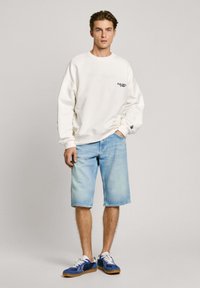Sudadera blanca con puños acanalados, combinada con pantalones cortos de mezclilla azul claro. El modelo lleva zapatillas azules y blancas. Diseño simple, corte casual.